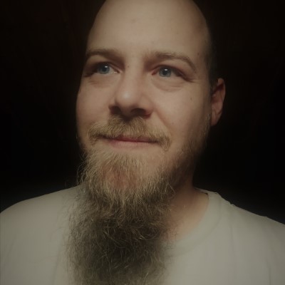 A young caucasian male shown from the shoulders up. He has blue eyes, a long blond beard and a light grey shirt.