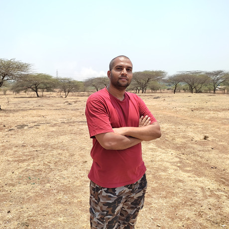 Onkar standing in the bushveld wearing a red short-sleeve shirt and long camo-pants.
