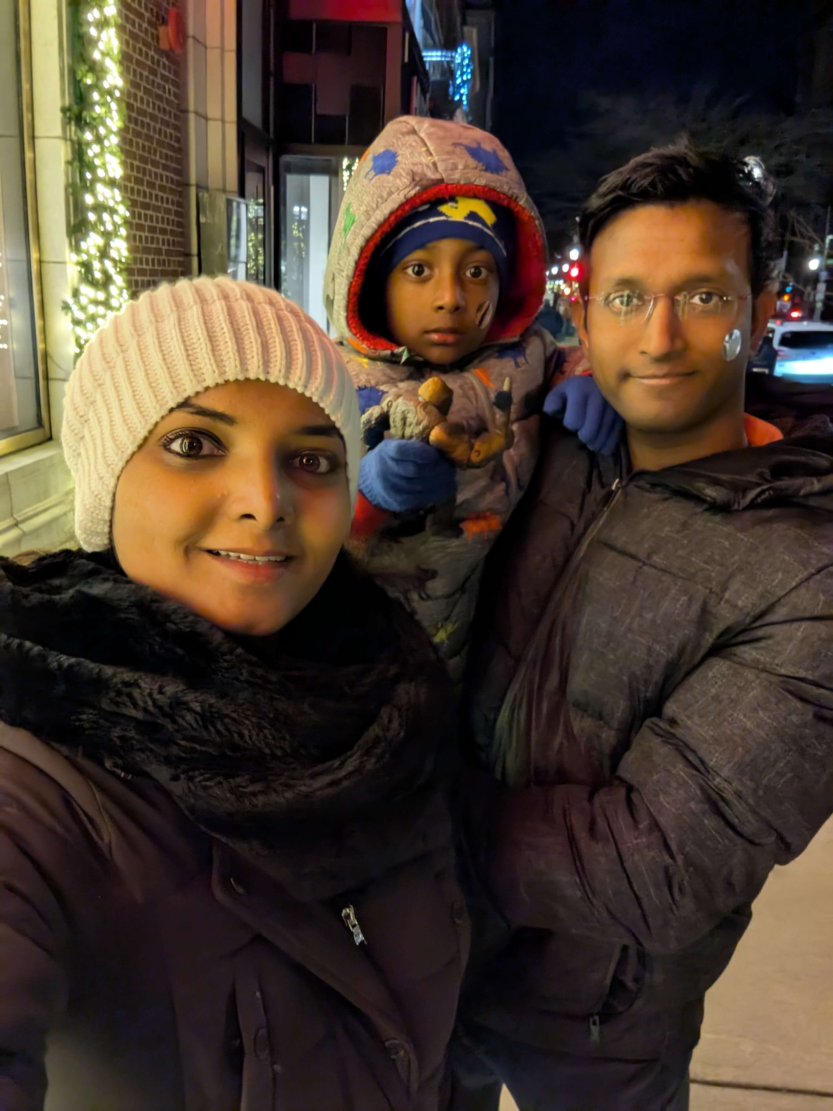 A family selfie taken at night on a city street decorated with holiday lights.  The street behind them is softly lit with festive lights and passing cars.