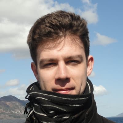 A young white male with short brown hair, wearing a scarf around his neck. Behind him is a mountain peak, with white clouds dotting a blue sky.